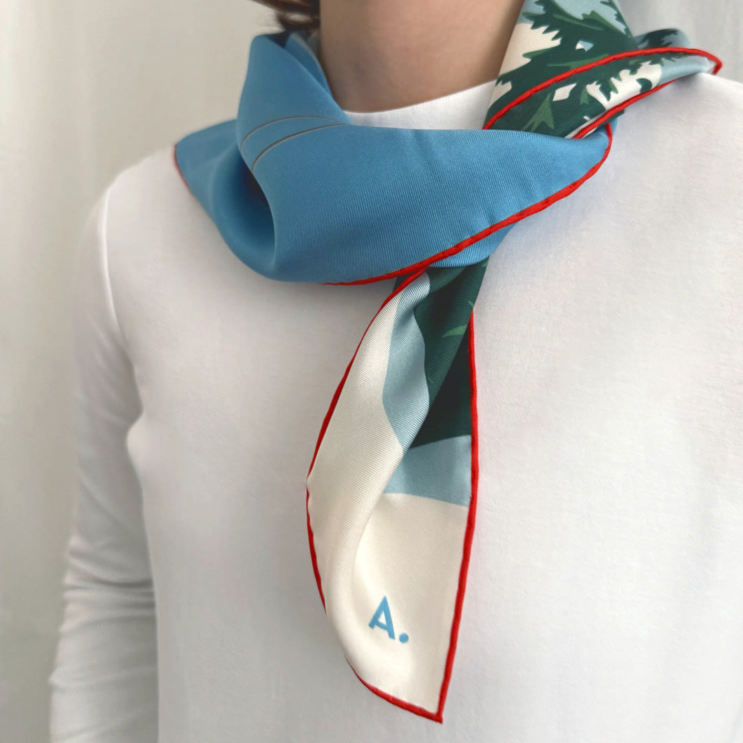 Frau mit einem Seidentuch um den Hals gebunden. Das Tuch zeigt eine Winterlandschaft in den Farben weiß, blau, Berge und Bäume und eine Gondel