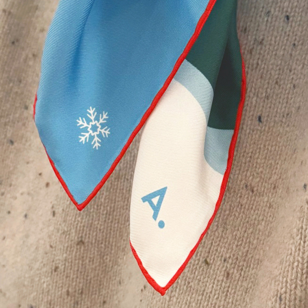 Frau mit einem Seidentuch um den Hals gebunden. Das Tuch zeigt eine Winterlandschaft in den Farben weiß, blau, Berge und Bäume und eine Gondel