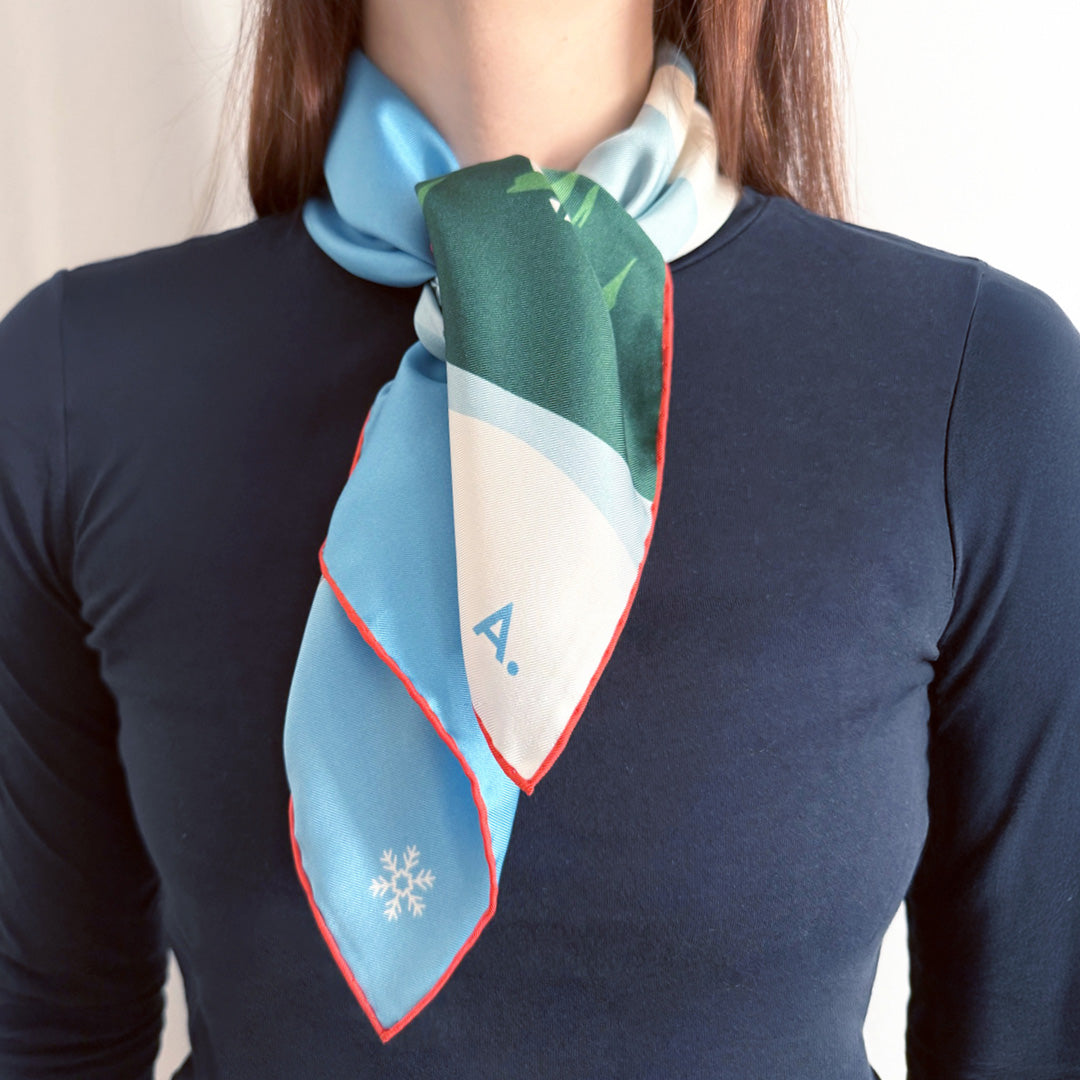 Frau mit einem Seidentuch um den Hals gebunden. Das Tuch zeigt eine Winterlandschaft in den Farben weiß, blau, Berge und Bäume und eine Gondel